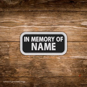 May include: A rectangular patch with rounded corners, featuring the text "IN MEMORY OF NAME" in white, centered on a black background. The patch has a white border and is set against a wooden background.