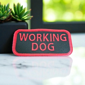 May include: A rectangular patch with the words "WORKING DOG" embroidered in red on a black background. The patch has a red border and is likely designed to be sewn onto a garment or accessory.
