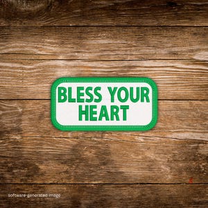May include: A rectangular patch with rounded corners, featuring the phrase "BLESS YOUR HEART" in bold green letters on a white background. The patch is outlined with a green border and rests on a wooden surface.