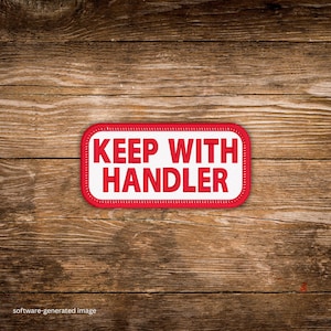 May include: A rectangular patch with the words "KEEP WITH HANDLER" in red block letters on a white background. The patch has a red border and is set against a wooden background.