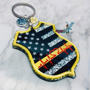 May include: A black and silver glitter resin keychain shaped like a police badge with a thin yellow line and a red heart with a heartbeat line. The keychain has a silver charm with the words "Love You" on it.