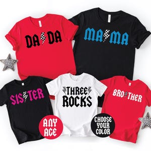 May include: A collection of five short-sleeved t-shirts in red, black, and white. The shirts feature graphic text designs including DADA, MAMA, SISTER, THREE ROCKS, and BROTHER, with checkerboard lightning bolt accents and circular text labels.
