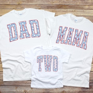 May include: Three white t-shirts with text. The largest shirt says "DAD" in blue gingham letters with red outlines. The second shirt says "MAMA" in the same style. The smallest shirt says "TWO".
