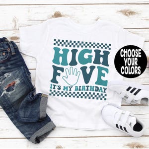 May include: A white t-shirt featuring a teal and dark teal graphic with a checkered border. The text reads HIGH FIVE IT'S MY BIRTHDAY, with a hand outline replacing the letter I. A black circle with the white text CHOOSE YOUR COLORS is included.