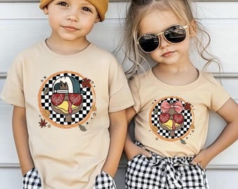May include: Two beige short-sleeve t-shirts featuring a graphic of a turkey wearing heart-shaped glasses with the text GOBBLE GOBBLE inside a black and white checkered circle, accented with autumn leaves and stars, worn with black and white gingham shorts.