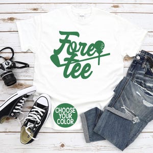 May include: White t-shirt with the words "Fore Tee" in green, along with a golf ball and tee design. The shirt is paired with blue jeans, black sneakers, and a camera. A green circle says "Choose Your Color".