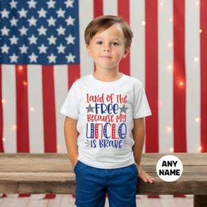 May include: A white short-sleeved t-shirt featuring the red, white, and blue text Land of The FREE Because My UNCLE is BRAVE with two grey stars. The shirt is paired with solid blue shorts, creating a patriotic look for a young child.