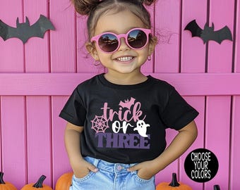 May include: A black short-sleeved t-shirt featuring a graphic print with the text "trick or THREE" in pink and purple, accompanied by a pink spiderweb, a small pink bat, and a white ghost. The shirt is paired with light blue denim shorts.