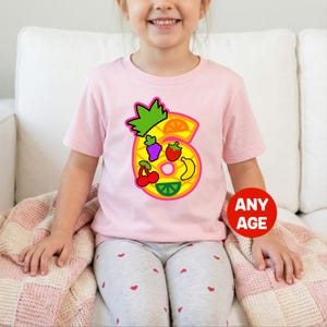 May include: A light pink short-sleeved t-shirt featuring a large graphic of the number six. The number is filled with a yellow pineapple pattern and decorated with colorful illustrations of orange slices, grapes, strawberries, bananas, cherries, and lime wedges. A red circle with the text ANY AGE is visible.