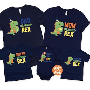 May include: Navy blue t-shirts and a onesie with dinosaur graphics and text. The shirts say "Dad", "Mom", "Sister", and "Brother" with "-osaurus- Rex" below. The onesie says "Roar I'm a One A-saurus".