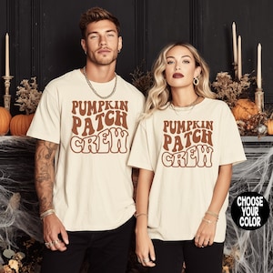 May include: Two people wearing cream-colored t-shirts with the text "PUMPKIN PATCH CREW" in brown, retro-style lettering. The shirts are short-sleeved and appear to be made of a soft material. The background includes Halloween decorations.