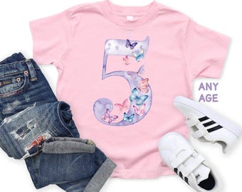 May include: A light pink short-sleeved t-shirt featuring a large purple watercolor-style number five decorated with various colorful butterflies. The shirt includes the text ANY AGE in purple. Also shown are distressed blue jeans and white sneakers.