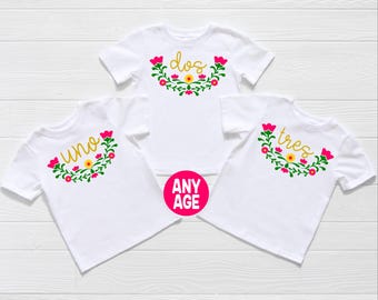 May include: Three white short-sleeved t-shirts featuring floral wreaths in green, pink, and yellow. Each shirt displays a cursive word in gold: uno, dos, or tres. A bright pink circular graphic with the white text ANY AGE is positioned below.