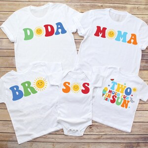 May include: A collection of five white t-shirts and one white baby bodysuit featuring colorful graphic text and sun illustrations. The shirts display the words DODA, MOMA, BROS, SOS, and TWO MUCH FUN IN THE SUN with various icons like ice cream.