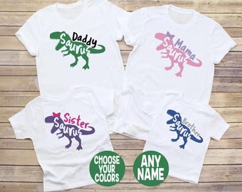 May include: Four white t-shirts featuring dinosaur graphics and text. The shirts display the words Daddy Saurus in green, Mama Saurus in pink, Sister Saurus in purple, and Brother Saurus in blue. Two green circles contain the text CHOOSE YOUR COLORS and ANY NAME.