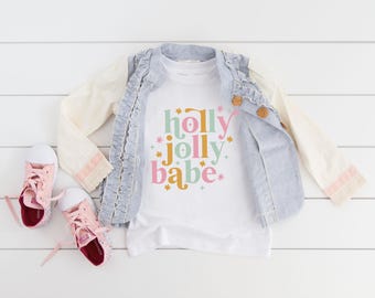 May include: A white t-shirt featuring the text "holly jolly babe" in pink, green, and gold, accented with small star shapes. The shirt is layered under a light blue ruffled vest with wooden buttons and paired with light pink canvas sneakers.