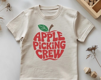 May include: A beige short-sleeved t-shirt featuring a large red graphic of an apple shape containing the text "APPLE PICKING CREW" in a wavy font, with a small green leaf and brown stem detail at the top of the apple design on the front.