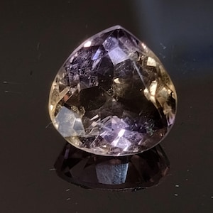 May include: A pear-shaped faceted gemstone displaying a blend of translucent purple and golden-yellow hues. The stone features numerous geometric cuts that create internal light reflections and a sharp pointed top with a rounded base.