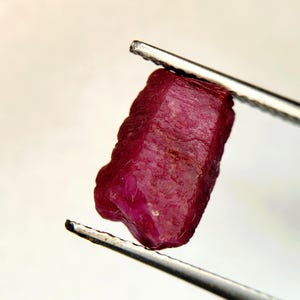 May include: A raw, deep red ruby gemstone held by silver-toned metal tweezers. The stone features a natural, unpolished, and textured surface with visible crystalline facets and irregular edges, showcasing a rich, saturated crimson color throughout.
