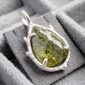 Peut inclure: Pendentif en argent avec une pierre précieuse verte olive en forme de poire et facettée. Le cadre du pendentif a un design texturé et organique, ressemblant à des branches. Le bijou est présenté sur une surface grise texturée.