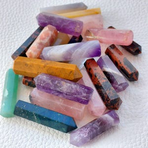 May include: A collection of various gemstone wands in shades of purple, pink, orange, green, and blue. The wands have a hexagonal shape and are polished to a smooth finish. The gemstones are arranged on a white surface.
