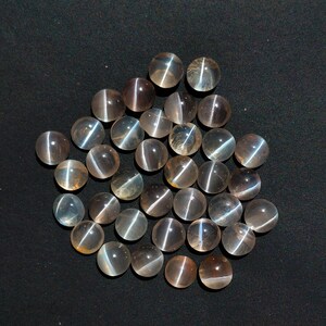 Puede incluir: Una colección de piedras preciosas redondas y pulidas con efecto ojo de gato. Las piedras son una mezcla de translúcidas y opacas, con una gama de colores que va del claro al marrón. Cada piedra tiene una línea brillante y distintiva en su centro. Las piedras están dispersas sobre una superficie negra.