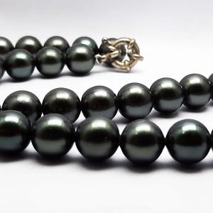 Puede incluir: Un collar de perlas negras con un cierre de plata. Las perlas son redondas y lisas, y están enhebradas en una fina cadena de plata.