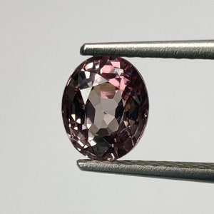 以下が含まれることがあります： ピンセットで保持された、丸くてファセットカットされたピンクの宝石。宝石は紫がかった色合いをしており、半透明です。