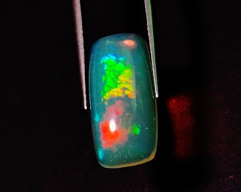 May include: A rectangular, polished opal gemstone with a dark teal base color. The opal displays vibrant internal colors of green, yellow, red, and blue. The gemstone is held by two silver metal prongs against a black background.