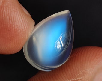Pode incluir: Um cabochão de pedra da lua em forma de lágrima, iridescente com um brilho azul. A pedra é segurada entre dois dedos.