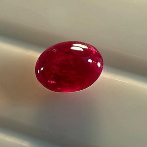 Può includere: Una gemma di forma ovale color rosso intenso con una superficie cabochon liscia e lucida. La pietra presenta una qualità traslucida con inclusioni interne e riflessi di luce bianca brillante sulla parte superiore curva, mostrando una ricca tonalità cremisi.