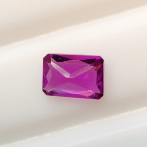 May include: A vibrant purple gemstone with a rectangular cut and a faceted finish. The gemstone is set against a white background.