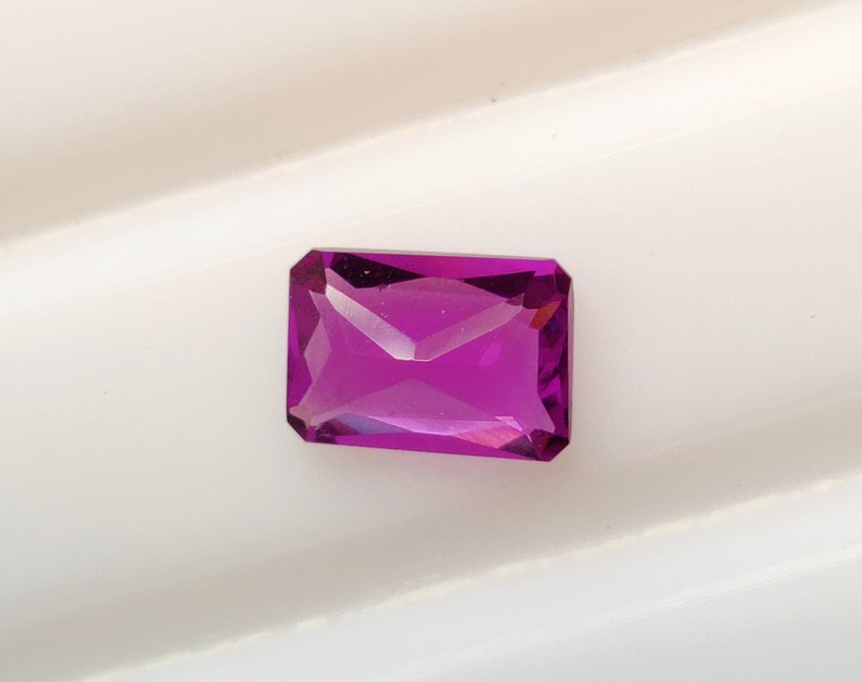 May include: A vibrant purple gemstone with a rectangular cut and a faceted finish. The gemstone is set against a white background.