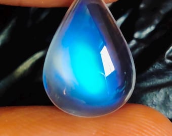 Puede incluir: Una piedra preciosa de piedra lunar en forma de pera con una superficie lisa y translúcida que presenta una prominente adularescencia azul brillante centrada. La gema muestra un acabado pulido y una silueta de lágrima.
