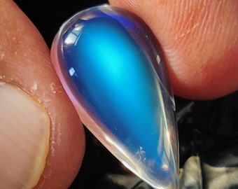 May include: A teardrop-shaped, iridescent gemstone with a vibrant blue center and clear edges. The polished stone reflects light, creating a shimmering effect. The gem is held between two fingers, showcasing its size and shape.