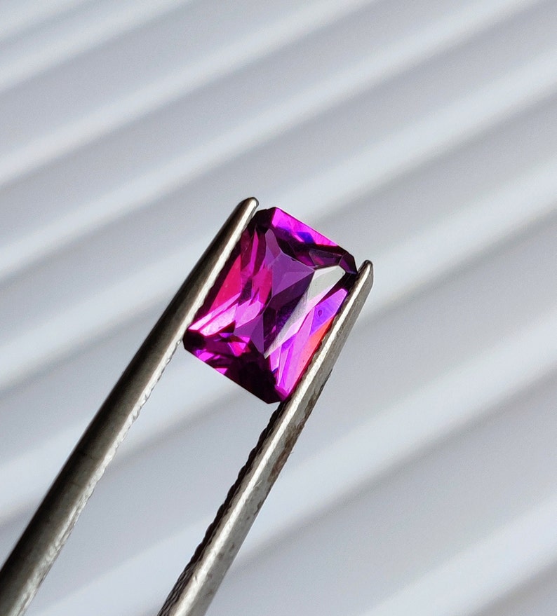 May include: A vibrant purple gemstone with a rectangular cut, held in tweezers against a white background.