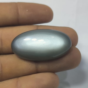 Puede incluir: Una gema gris, lisa y de forma ovalada. La superficie pulida refleja la luz, creando reflejos. La piedra se sostiene en una mano, mostrando su tamaño y forma. Esta gema podría usarse en joyería.