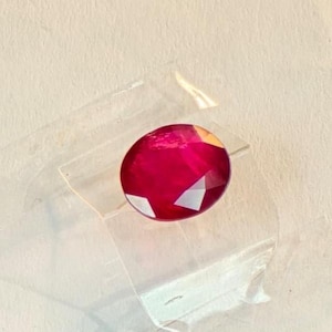 Puede incluir: Una gema de rubí ovalada y facetada. La gema es de un color rojo intenso y vibrante con múltiples facetas que reflejan la luz. El rubí está sobre una superficie clara y transparente.