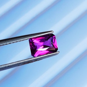 May include: A vibrant purple gemstone with a rectangular cut, held in tweezers against a blue and white striped background.