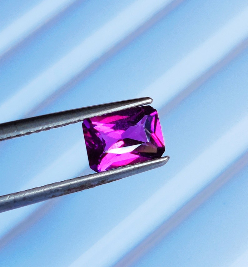 May include: A vibrant purple gemstone with a rectangular cut, held in tweezers against a blue and white striped background.