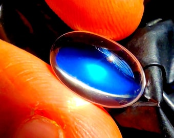 Pode incluir: Uma pedra preciosa em formato de cabochão oval com um centro azul profundo e translúcido que transita para um tom ciano mais claro e brilhante. A pedra possui uma superfície lisa e polida com qualidade reflexiva e luz interna visível.
