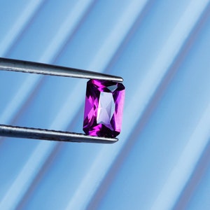 May include: A purple gemstone with a rectangular shape is held by tweezers against a blue background.