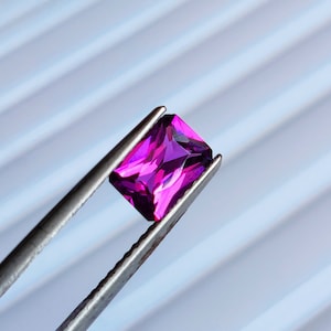 May include: A vibrant purple gemstone, likely amethyst, is held by tweezers against a white striped background. The gemstone is cut into a rectangular shape with facets that catch the light.