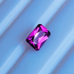 May include: A vibrant, deep purple gemstone with a rectangular cut and a faceted finish. The gemstone is set against a white background.