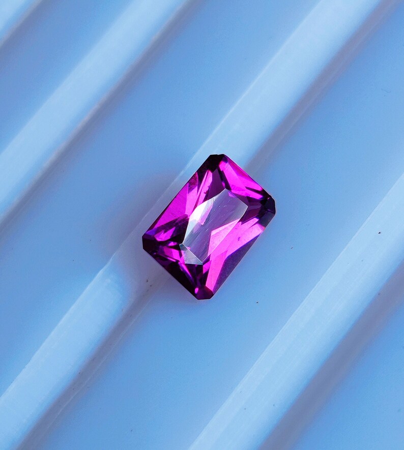 May include: A vibrant, deep purple gemstone with a rectangular cut and a faceted finish. The gemstone is set against a white background.