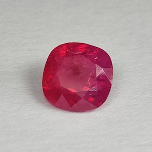 Puede incluir: Una gema roja vibrante con un corte cojín. La gema está facetada y tiene un alto pulido.