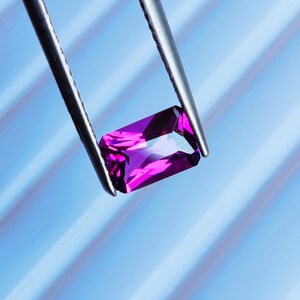 May include: A vibrant purple gemstone held by tweezers against a blue striped background.
