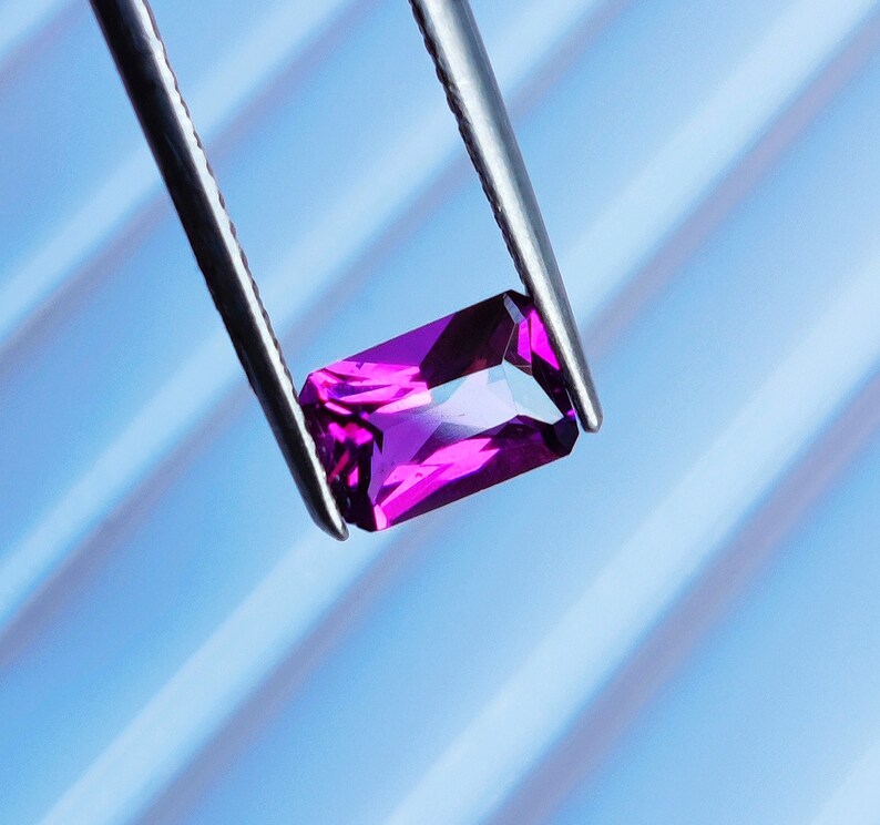 May include: A vibrant purple gemstone held by tweezers against a blue striped background.