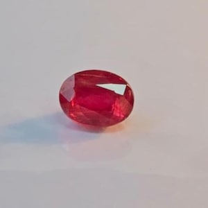 Puede incluir: Una gema de rubí facetada, de forma ovalada. La gema es de un color rojo intenso y saturado, con inclusiones internas visibles y un alto grado de claridad. Las superficies pulidas reflejan la luz.