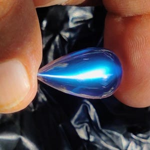 Puede incluir: Una gema azul iridiscente en forma de lágrima. La gema se sostiene entre dos dedos, mostrando su superficie pulida y la refracción interna de la luz. El fondo es un material oscuro y texturizado.